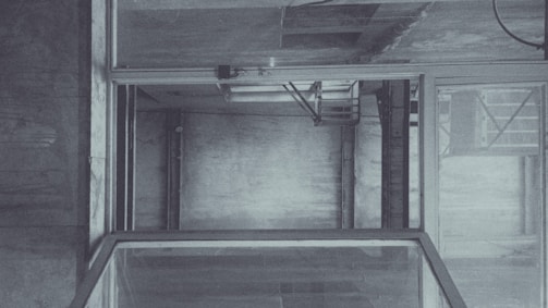 An empty, industrial elevator shaft with visible metal beams and concrete walls. The structure is framed by glass panels and has a utilitarian design. Subtle lighting casts soft shadows, adding depth to the interior.