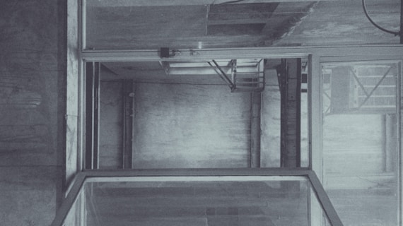 An empty, industrial elevator shaft with visible metal beams and concrete walls. The structure is framed by glass panels and has a utilitarian design. Subtle lighting casts soft shadows, adding depth to the interior.