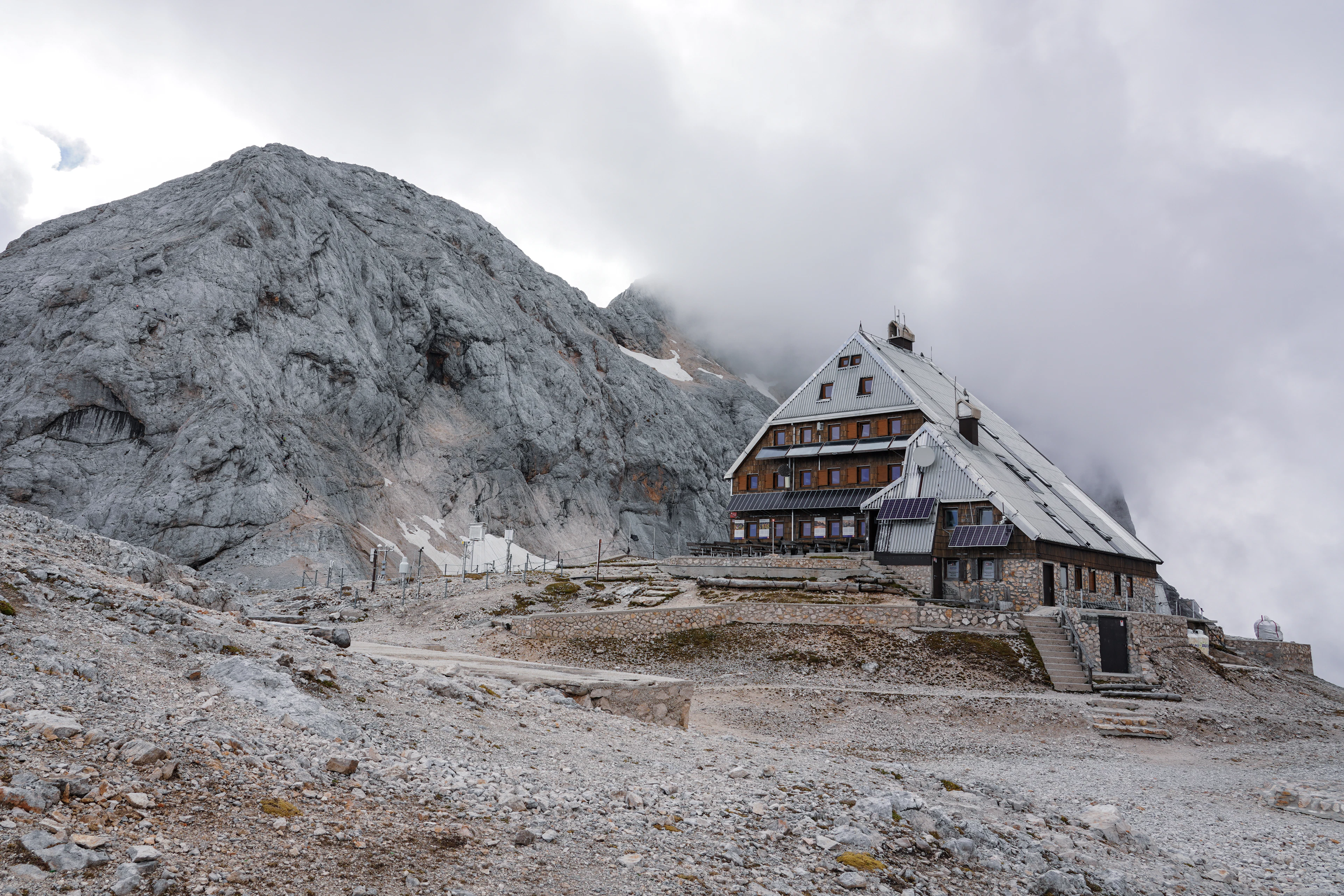 A large stone mountain hut among grey rocks, with a limestone massif rising into cloud above