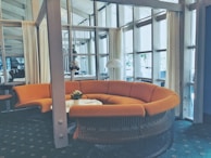 A modern interior space featuring a large, curved orange sofa set against a backdrop of tall windows with sheer curtains. A small round table sits in front of the sofa, adorned with a potted plant. The room has a contemporary design, with glass walls and a view of another room with tables and chairs.