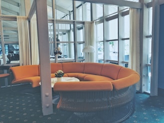 A modern interior space featuring a large, curved orange sofa set against a backdrop of tall windows with sheer curtains. A small round table sits in front of the sofa, adorned with a potted plant. The room has a contemporary design, with glass walls and a view of another room with tables and chairs.