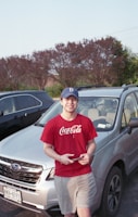 A young person wearing a red Coca-Cola T-shirt and a blue cap stands in front of a silver Subaru car in a parking lot. The person is smiling while holding a smartphone. In the background, there are several parked cars and a row of trees.
