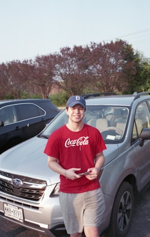 A young person wearing a red Coca-Cola T-shirt and a blue cap stands in front of a silver Subaru car in a parking lot. The person is smiling while holding a smartphone. In the background, there are several parked cars and a row of trees.