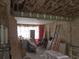 A room under renovation with exposed wooden beams and insulation on the ceiling, a bare brick wall, and various construction materials scattered around. There is a metal ladder leaning against the wall, a stack of drywall sheets, and bags of cement on the floor. The back of the room is illuminated by a large window covered with white curtains, with a red brick wall to the right.