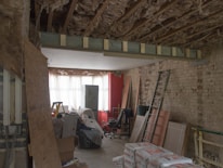 A room under renovation with exposed wooden beams and insulation on the ceiling, a bare brick wall, and various construction materials scattered around. There is a metal ladder leaning against the wall, a stack of drywall sheets, and bags of cement on the floor. The back of the room is illuminated by a large window covered with white curtains, with a red brick wall to the right.