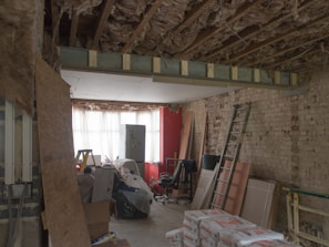 A room under renovation with exposed wooden beams and insulation on the ceiling, a bare brick wall, and various construction materials scattered around. There is a metal ladder leaning against the wall, a stack of drywall sheets, and bags of cement on the floor. The back of the room is illuminated by a large window covered with white curtains, with a red brick wall to the right.