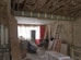 A room under renovation with exposed wooden beams and insulation on the ceiling, a bare brick wall, and various construction materials scattered around. There is a metal ladder leaning against the wall, a stack of drywall sheets, and bags of cement on the floor. The back of the room is illuminated by a large window covered with white curtains, with a red brick wall to the right.