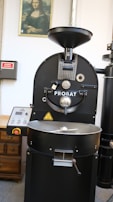 A large industrial coffee roasting machine with the brand name Probat is positioned centrally. Above the machine is a framed picture of the Mona Lisa on the wall. To the left, there's a wooden cabinet and an electrical panel with a red sign. The setting appears to be a coffee processing facility or similar environment.