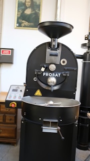 A large industrial coffee roasting machine with the brand name Probat is positioned centrally. Above the machine is a framed picture of the Mona Lisa on the wall. To the left, there's a wooden cabinet and an electrical panel with a red sign. The setting appears to be a coffee processing facility or similar environment.
