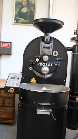 A large industrial coffee roasting machine with the brand name Probat is positioned centrally. Above the machine is a framed picture of the Mona Lisa on the wall. To the left, there's a wooden cabinet and an electrical panel with a red sign. The setting appears to be a coffee processing facility or similar environment.