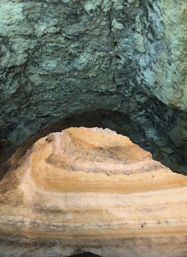 An image showing the interior of a cave with textured, stratified rock formations. The layers of rock vary in color from dark grey to a lighter sand-like hue, indicating different mineral compositions and erosion patterns.