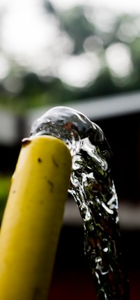 A stream of clear water flows smoothly from the opening of a yellow garden hose, with a blurred natural background suggesting an outdoor setting.