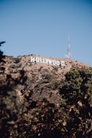 A hillside landscape featuring the iconic Hollywood sign against a clear blue sky. The surrounding area has sparse vegetation, and the terrain includes rocky and dry sections. A radio tower is visible near the sign, adding an industrial element to the natural scene.