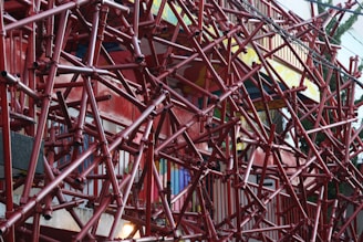 An intricate network of red pipes and scaffolding covering a building facade. The pipes create a complex geometric pattern, giving the structure an industrial and chaotic look. The background features colorful walls and railings, partially visible through the scaffolding.