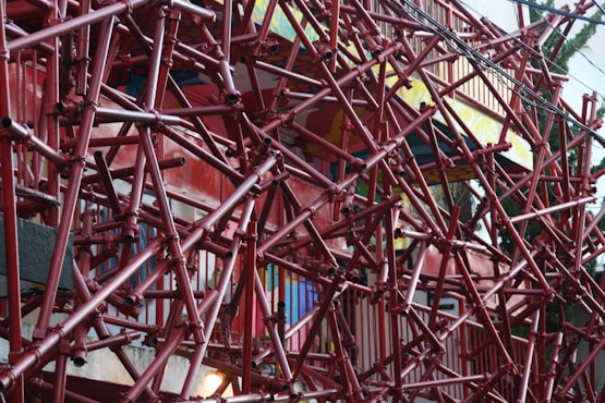 An intricate network of red pipes and scaffolding covering a building facade. The pipes create a complex geometric pattern, giving the structure an industrial and chaotic look. The background features colorful walls and railings, partially visible through the scaffolding.