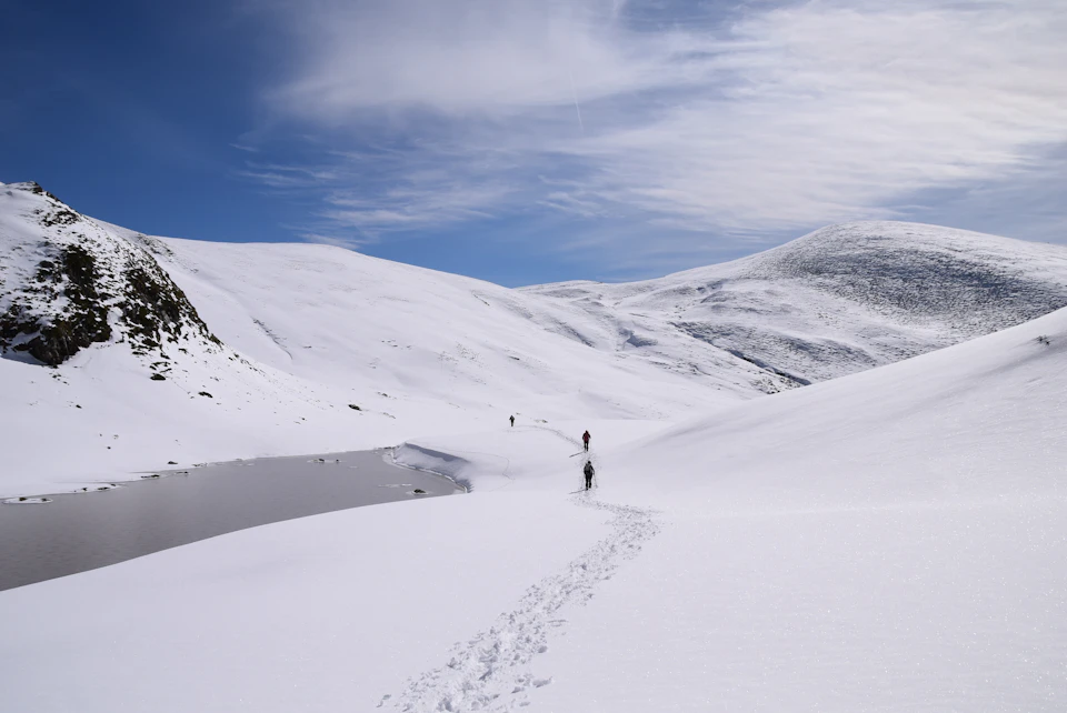 The image depicts a snowy mountain landscape with two hikers making their way up the side of a steep, snow-covered slope. The hiker on the left is slightly ahead of the other, who is following closely behind. Both hikers are wearing backpacks and appear to be in good physical condition for the challenging hike. They are moving towards the right side of the image, leaving footprints in the snow as they ascend the mountain. In the background, a small lake can be seen nestled among the snowy mountains, adding depth and tranquility to the scene.