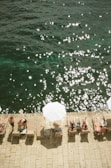 People are relaxing on sun loungers by the water's edge, with sun umbrellas providing shade. The water is sparkling under the sunlight, creating a serene and summery atmosphere. The scene captures leisure and tranquility in a coastal setting.