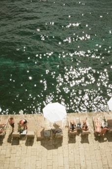 People are relaxing on sun loungers by the water's edge, with sun umbrellas providing shade. The water is sparkling under the sunlight, creating a serene and summery atmosphere. The scene captures leisure and tranquility in a coastal setting.
