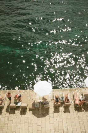 People are relaxing on sun loungers by the water's edge, with sun umbrellas providing shade. The water is sparkling under the sunlight, creating a serene and summery atmosphere. The scene captures leisure and tranquility in a coastal setting.