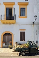 A small three-wheeled vehicle parked in front of a white building with yellow accents. The vehicle's flatbed is filled with various potted plants. The building features two windows with black frames and a small balcony above a wooden doorway. Two potted plants are positioned on either side of the door, adding to the rustic charm.