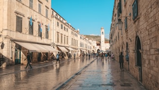 A historic street lined with stone buildings features a wet cobblestone path with people walking and exploring the area. Flags hang on some of the buildings, and a prominent clock tower stands in the distance, adding to the architectural appeal of the scene. The sky is clear and blue, suggesting a pleasant day.
