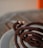 A coiled, dark brown incense stick is burning, with a wisp of smoke rising from its tip. It rests on a striped plate, and the background is softly blurred, featuring warm tones.