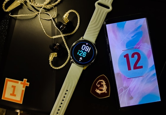 A smartwatch with a white band displays the time and date. Next to it is a smartphone showing the number 12 on a colorful screen. Earphones with a braided wire are placed nearby alongside a black object with a logo resembling a dragon. A red and white logo sits partly off-frame on a black surface.