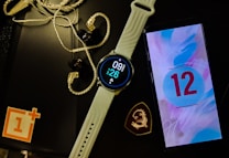 A smartwatch with a white band displays the time and date. Next to it is a smartphone showing the number 12 on a colorful screen. Earphones with a braided wire are placed nearby alongside a black object with a logo resembling a dragon. A red and white logo sits partly off-frame on a black surface.