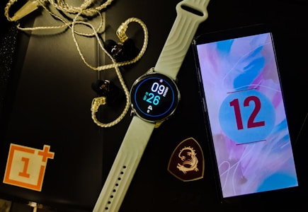 A smartwatch with a white band displays the time and date. Next to it is a smartphone showing the number 12 on a colorful screen. Earphones with a braided wire are placed nearby alongside a black object with a logo resembling a dragon. A red and white logo sits partly off-frame on a black surface.