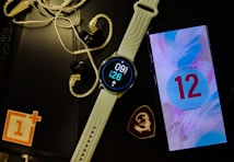 A smartwatch with a white band displays the time and date. Next to it is a smartphone showing the number 12 on a colorful screen. Earphones with a braided wire are placed nearby alongside a black object with a logo resembling a dragon. A red and white logo sits partly off-frame on a black surface.
