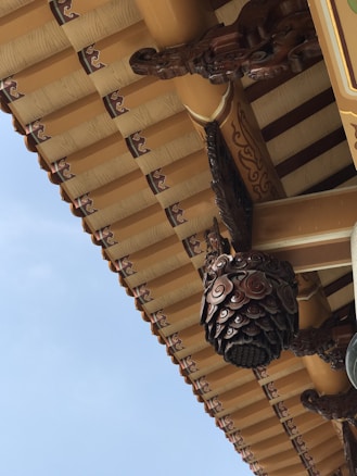 A traditional architectural roof structure features intricate wooden carvings and design elements beneath a series of curved, tiled eaves. The ornate patterns and details suggest historical or cultural significance in the construction. The image is taken from below the structure, highlighting the decorative craftsmanship and the clear blue sky in the background.