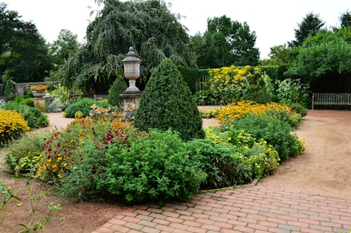 A lush, well-maintained garden with a variety of flowering plants in full bloom. The garden features neatly trimmed hedges, vibrant flowers in shades of yellow, red, and orange, and a stone urn on a pedestal. Gravel paths and a brick walkway intersect the garden, with trees and shrubs providing a rich green backdrop.