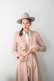 A person stands wearing a stylish beige trench coat with a matching beige hat. The person has a hairstyle with a side braid and sports bright red lipstick. The background is a plain light tone, enhancing the overall minimalist and chic aesthetic of the outfit.