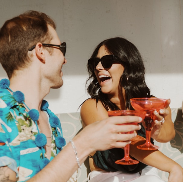 A man and a woman are sitting closely together, both wearing sunglasses. They are engaged in a cheerful moment, holding red cocktail glasses and smiling warmly at each other. The woman has long, dark hair, while the man is wearing a colorful shirt with a floral pattern and a blue lei around his neck.