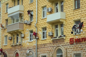 Workers are suspended by ropes on the side of a yellow geometric-patterned building, painting or repairing the facade. The building features multiple balconies and air conditioning units attached to the walls. The lower part of the building has a textured stone appearance, and there's a red sign with Cyrillic lettering.