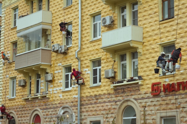 Workers are suspended by ropes on the side of a yellow geometric-patterned building, painting or repairing the facade. The building features multiple balconies and air conditioning units attached to the walls. The lower part of the building has a textured stone appearance, and there's a red sign with Cyrillic lettering.