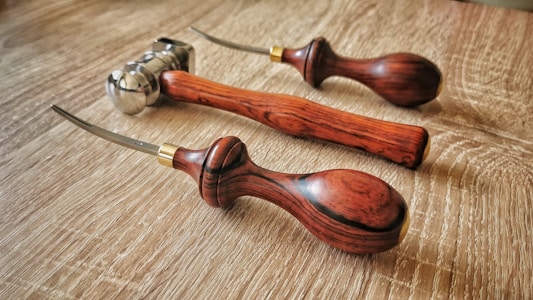 Three leatherworking tools with wooden handles rest on a wooden surface. The tools have curved metal tips and brass accents near the handles.