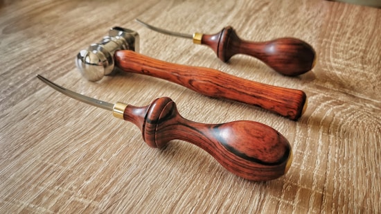 Three leatherworking tools with wooden handles rest on a wooden surface. The tools have curved metal tips and brass accents near the handles.