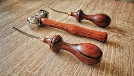 Three leatherworking tools with wooden handles rest on a wooden surface. The tools have curved metal tips and brass accents near the handles.
