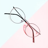 A pair of round eyeglasses is placed on a background divided diagonally into two pastel-colored sections, light blue and pale pink. The metal frame of the glasses contrasts sharply with the softly hued backdrop.