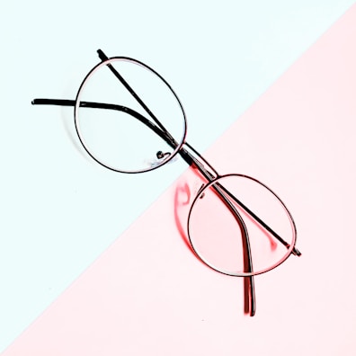A pair of round eyeglasses is placed on a background divided diagonally into two pastel-colored sections, light blue and pale pink. The metal frame of the glasses contrasts sharply with the softly hued backdrop.