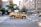 A yellow taxi is driving quickly through an urban street. The background shows blurred buildings and trees, indicating motion and speed.