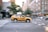 A yellow taxi is driving quickly through an urban street. The background shows blurred buildings and trees, indicating motion and speed.
