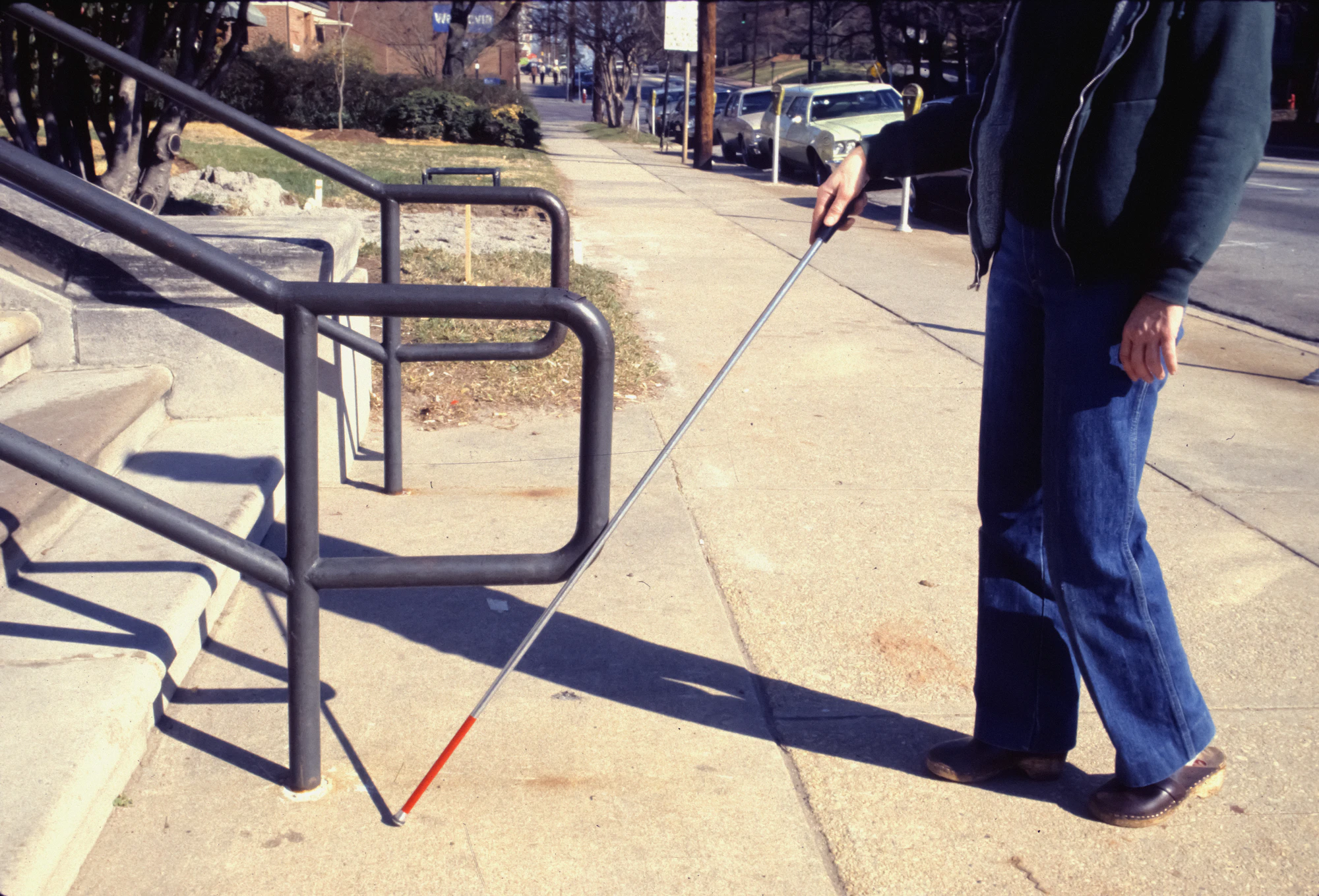 A man walking along a pavement with a long white cane