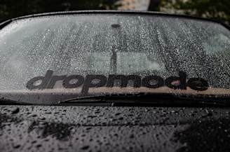 Raindrops covering a car windshield, with a visible sticker displaying the word 'dropmode'. The background is slightly blurred, hinting at a tree and possibly a building.