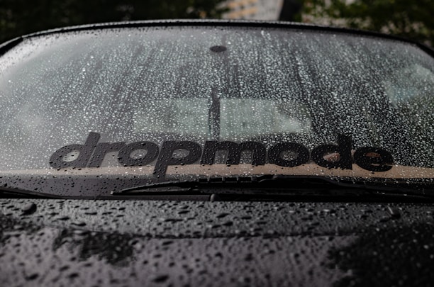 Raindrops covering a car windshield, with a visible sticker displaying the word 'dropmode'. The background is slightly blurred, hinting at a tree and possibly a building.