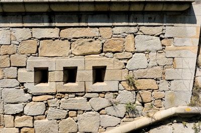 A stone wall constructed from large, rough-hewn blocks and mortar with three small, square openings arranged horizontally. Some greenery, such as small plants or moss, grows between the stones.