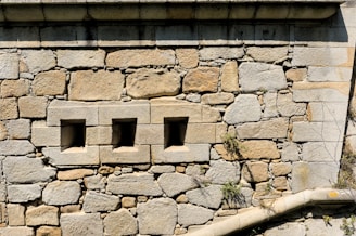A stone wall constructed from large, rough-hewn blocks and mortar with three small, square openings arranged horizontally. Some greenery, such as small plants or moss, grows between the stones.