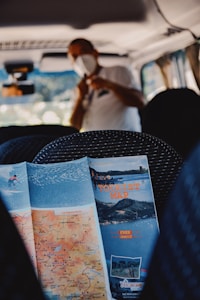 A map with the label 'TOURIST MAP' and images of landscapes is positioned on the passenger seat inside a vehicle. In the background, a person wearing a white shirt and a mask is slightly blurred, suggesting the interior setting of a van or bus with seating and subdued lighting.