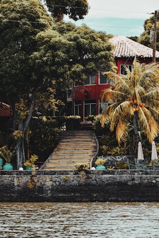 A picturesque house with red walls and a tiled roof is partially hidden behind lush green trees and palm trees. Stone steps lead up from a riverbank, with the house nestled among the foliage. A stone retaining wall borders the water, and there are green pots and outdoor furniture near the entrance.